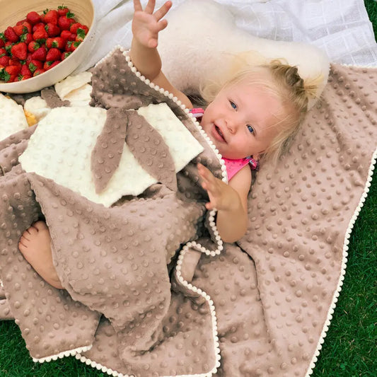 A baby lies on a plush blanket with a bowl of strawberries nearby. 2in1 Blanket with Baby Comforter in Cappuccino for sensory development and comfort.
