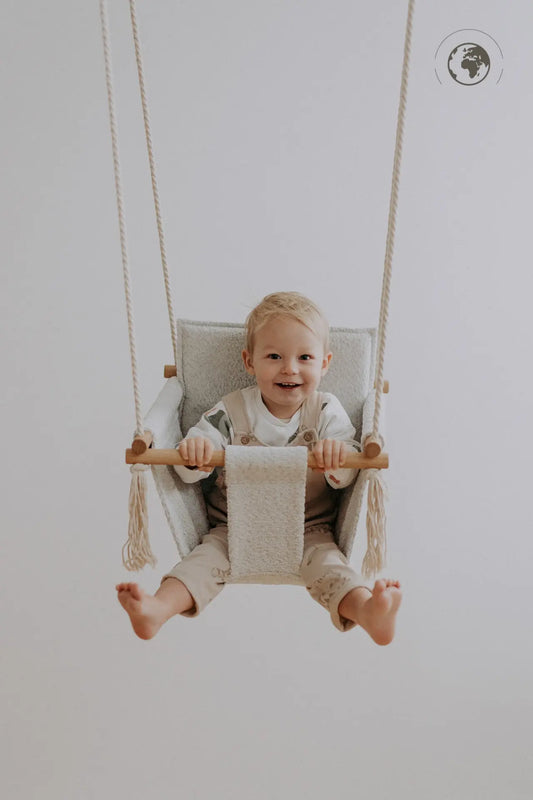 A baby happily swings in the luxurious Baby Swing - Bouclé Ice Grey, made from recycled materials and oak wood poles for safety and style.