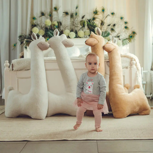 A baby stands among llamas, a plush toy alpaca on a bed, and a pair of llamas. Giant Plush Toy Alpaca in Beige, handmade with soft vegan fur, eco-friendly materials, and CE safety standards.