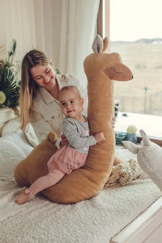 A woman holding a baby on a stuffed animal, emphasizing the cuddly comfort and whimsical charm of our Giant Plush Toy Alpaca in Rust.