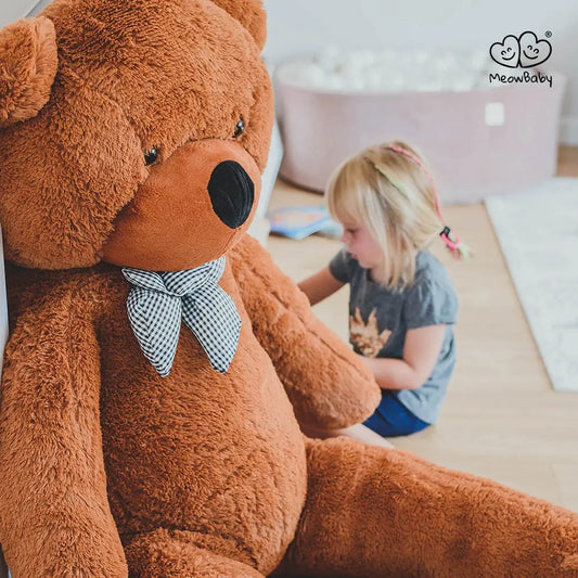 A girl sits next to a large teddy bear, embodying the Giant Teddy - 180 cm. Soft fur, EU safety certified, washable, and made for joyful hugs. Ideal for gifting smiles and cuddles.
