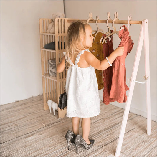 A girl in a white dress stands near a Kids Wooden Clothing Rack, showcasing its Montessori-style design with accessible shelves for easy outfit selection. Encourages independence and organization in children.