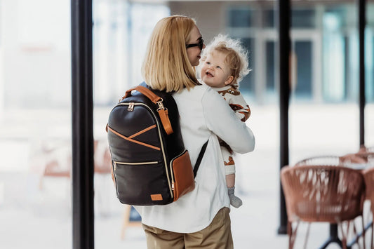 A woman carries a baby while wearing a black coffee diaper bag. Spacious with 16 pockets, laptop and baby food compartments, stroller straps, and a changing mat.
