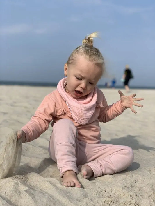 A child wearing a handmade double-sided muslin scarf-bib in light pink, sitting on sand. Crafted for style and protection from autumn breeze and saliva.