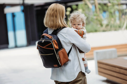 A woman carrying a baby in a Small Diaper Backpack – Black Coffee, designed for style and convenience. Features multiple pockets, stroller straps, and quality vegan leather.