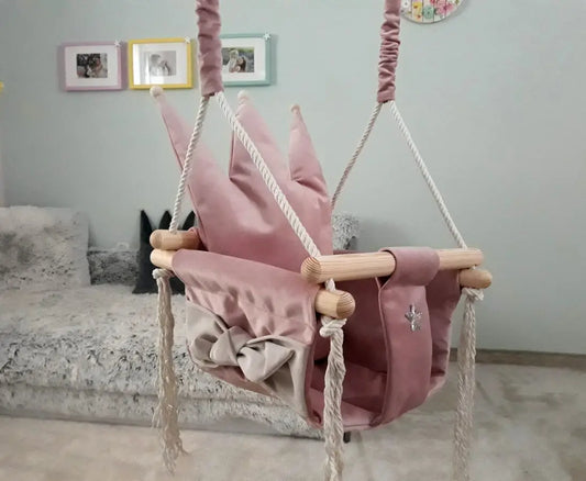 A pink swing with a crown-shaped cushion, crafted from velvet plush, featuring a podium crown for comfort. Made of durable pinewood, includes cotton ropes, and metal wheels for hanging.