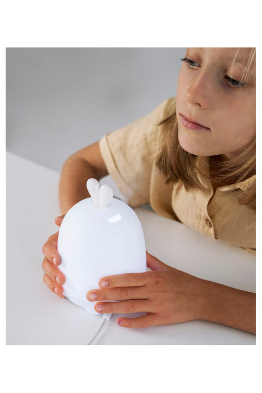 A young girl holds a white Rabbit Diffuser Lamp with bunny ears, emitting cool mist for air humidification and aromatherapy. LED light changes colors for night use, promoting healthy sleep.