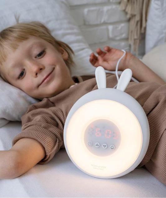 Child in bed with Rabbit Lamp alarm clock, sunrise/sunset simulation, nature sounds, touch control, snooze, and light intensity adjustment. Enhances sleep routines with gentle wake-up and bedtime signals. Dimensions: 19.3 x 15 x 6.9 cm.