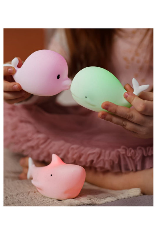 A girl holds two plastic whales, a hand holds a green balloon, and a pink toy. A waterproof set of 3 mini marine silicone lamps, perfect for kids' rooms and bath time.