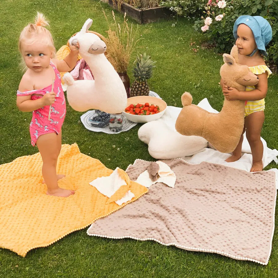 Two children on blankets in a grassy yard, one holding a candy cane and a stuffed animal. A sensory 2in1 Blanket with Baby Comforter in Cappuccino, featuring plush and satin fabrics for all seasons, textured bubbles for sensory stimulation, and a baby security toy with bunny ears.