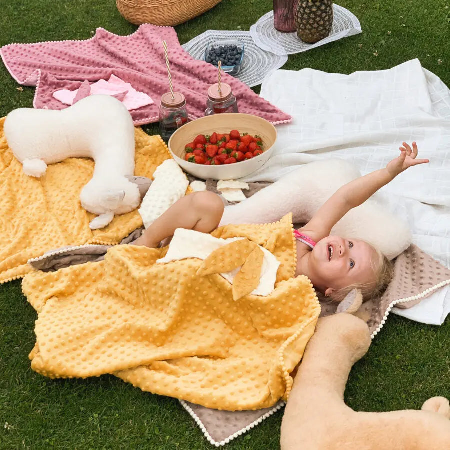 A girl lies on a plush blanket on grass, holding a bowl of strawberries. 2in1 Blanket with Baby Comforter in Mustard, designed for sensory development, warmth, and comfort.