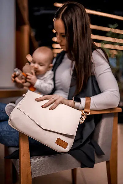 A woman holds a white handbag with a baby, showcasing the versatile Baby Accessories Handbag – Cappuccino. Features long and wrist straps for flexible carrying options.