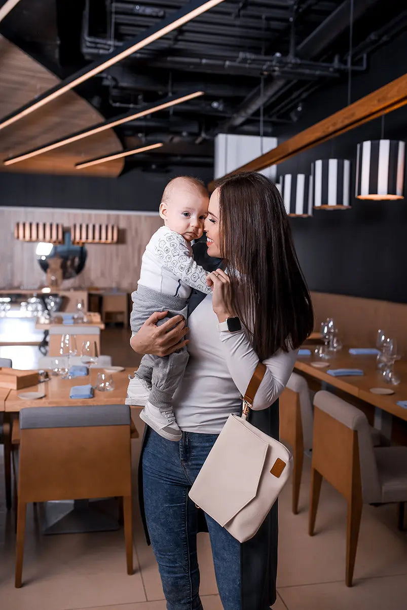 A woman holding a baby, showcasing the Baby Accessories Handbag – Cappuccino by Ally Scandic. Versatile design with long and wrist straps for convenient carrying.