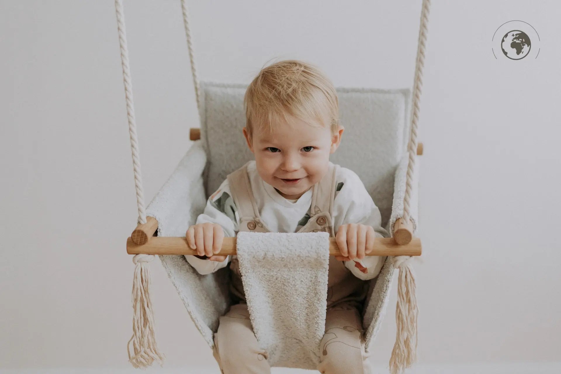 A baby happily swings in the luxurious Baby Swing - Bouclé Ice Grey, crafted from recycled materials for safety and comfort.