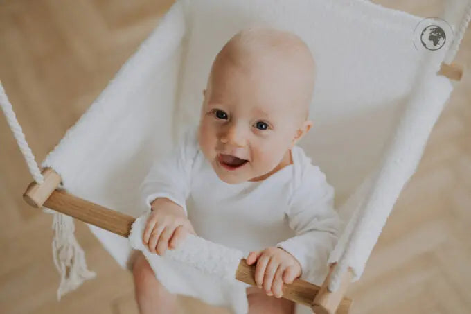 A baby in a white swing, made from recycled materials, oak wood poles, and bouclé cloth. Certified safe and stylishly Nordic, complete with pillow and carabiners. Dimensions: 41 x 41 cm.