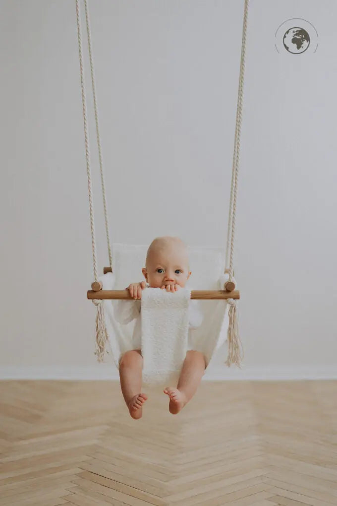 A baby happily swings in a luxurious Baby Swing - Bouclé Snow White made from recycled materials and oak wood poles, ensuring safety and comfort indoors.