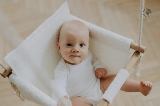 A baby in a white cloth swing, made from recycled materials and oak wood poles, certified for safety, easy to clean, and stylishly Nordic in design. Dimensions: 41 x 41 cm.