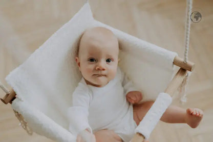 A baby in a white cloth swing, made from recycled materials and oak wood poles, certified for safety, easy to clean, and stylishly Nordic in design. Dimensions: 41 x 41 cm.