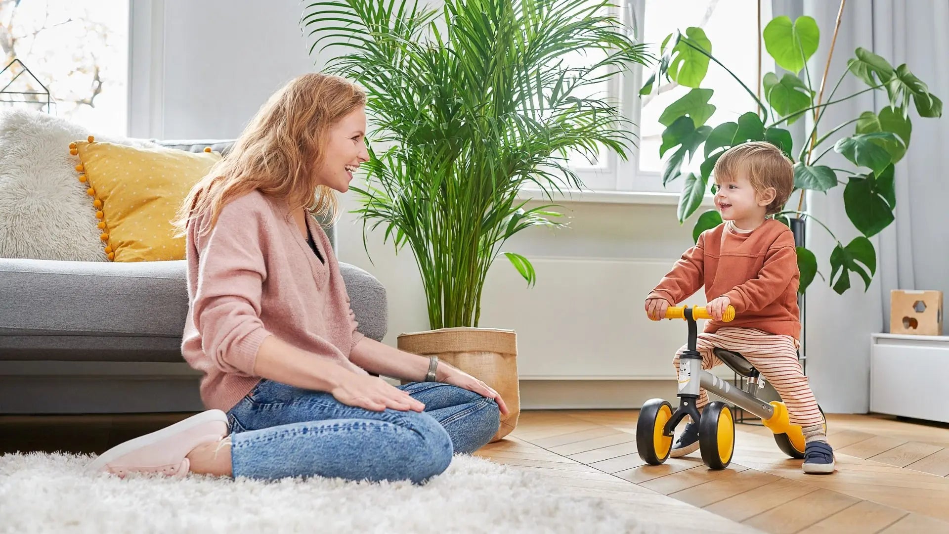 A woman and child enjoy the Balance Bike CUTIE - Yellow. Child on tricycle, woman sitting, plant nearby. Durable steel frame, adjustable saddle, rubber handles for safe play.