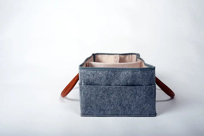 A grey felt Storage Basket / Diaper Caddy Organizer with brown vegan leather handles and multiple pockets for baby supplies. Lightweight, durable, and customizable for effortless parenting on-the-go.