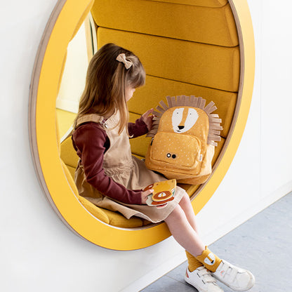 A girl sits in a circular object, holding a yellow backpack with a lion face. Backpack - Mr Lion: adjustable, water-repellent, 100% cotton, 7.5L capacity, ideal for ages 3+.