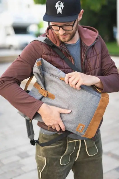 A man holding a grey and brown diaper backpack Berlin with magnetic closure, adjustable size, changing pad, bottle pocket, and padded straps.