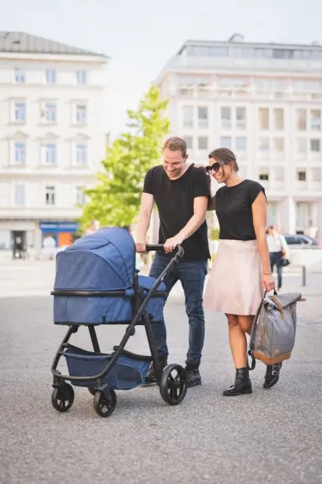 A man and woman push a stroller. Woman in a skirt. Man in black shirt. Baby essentials backpack Berlin with magnetic closure, adjustable size, reflective attachments, changing pad, bottle pocket, padded straps.