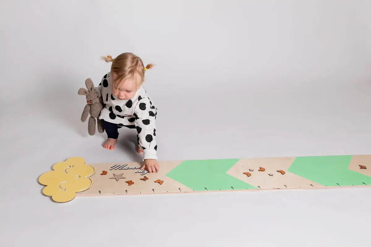 A toddler with a stuffed animal and a baby in a polka dot shirt, interacting with a Mint Growth Chart featuring a baby blue design with clouds and raindrops. Size: 27 x 154 cm.