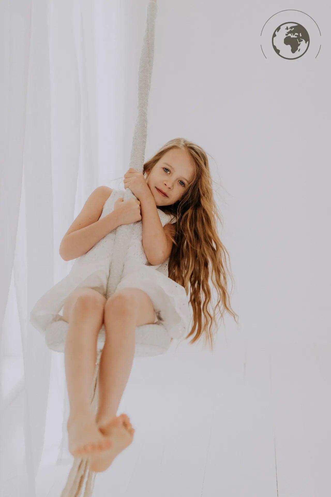 A girl in a white dress joyfully swings on the HopOn Swing - Bouclé Ice Grey, promoting physical development and pure joy. Dimensions: 31 cm seat, 2.5 m cotton rope, 100 kg capacity. OEKO-TEX certified materials.