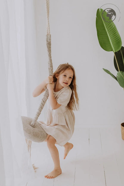 A girl joyfully swings on the HopOn Swing - Bouclé Sand, promoting physical development and pure joy. Fabric and cotton rope, OEKO-TEX certified, suitable for indoor use. Load capacity up to 100 kg.