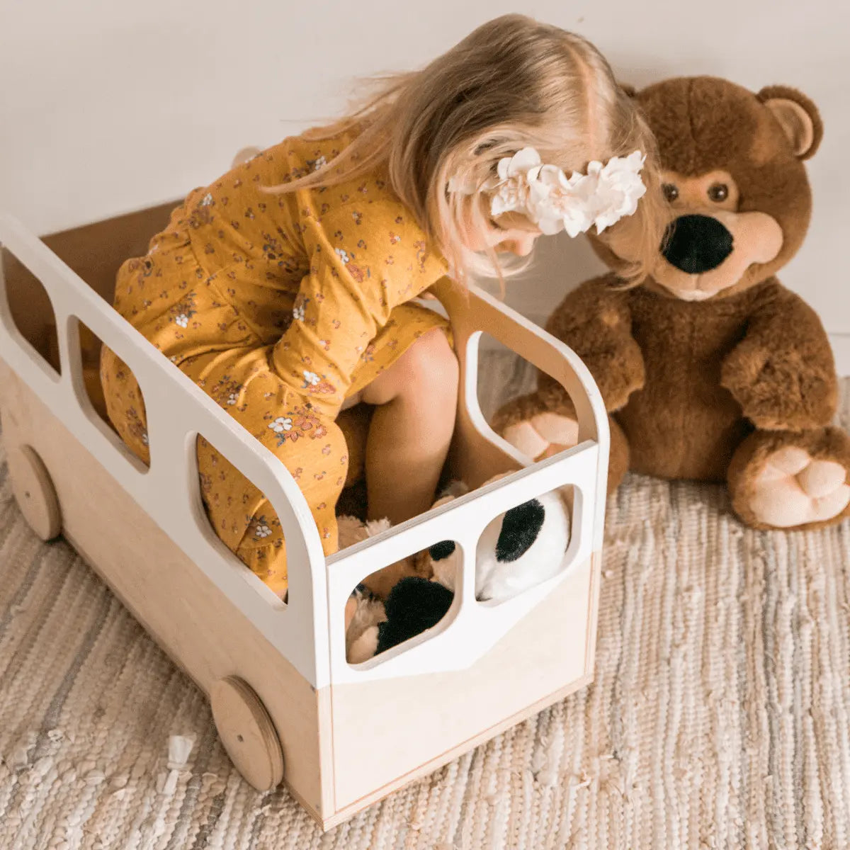 A girl sits in a toy car next to a teddy bear, embodying playful storage with our Kids Toy Storage Box on Wheels - Bus. Functional, mobile, and safe for children. Dimensions: 59 x 33 x 28 cm.