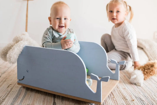 A baby sitting on a toy boat, a child sitting on a bed, and a baby holding a blue object. Product: Kids Toy Storage Box on Wheels - Whale. Dimensions: 48 x 33 x 23 cm. Functional design for easy toy organization. Safe plywood material.