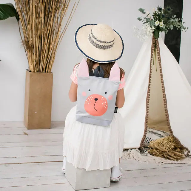 A girl sits on a box with a Bunny backpack, hat, and backpack. The backpack, made of velvet and polyester, features a Bunny design, adjustable straps, and a zipper closure. Ideal for 2-6-year-olds.