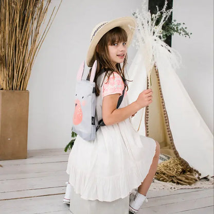 A girl in a white dress with a Bunny backpack, ready for adventures. Lightweight, velvet top, waterproof polyester bottom, adjustable straps. Ideal for 2-6-year-olds. Height: 30cm, Width: 26cm.