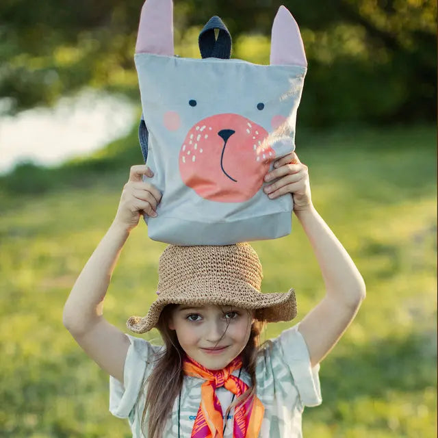 A young girl outdoors holds a Bunny kids backpack on her head. The backpack features a velvet top with a Bunny design and sturdy polyester bottom. Perfect for ages 2-6.