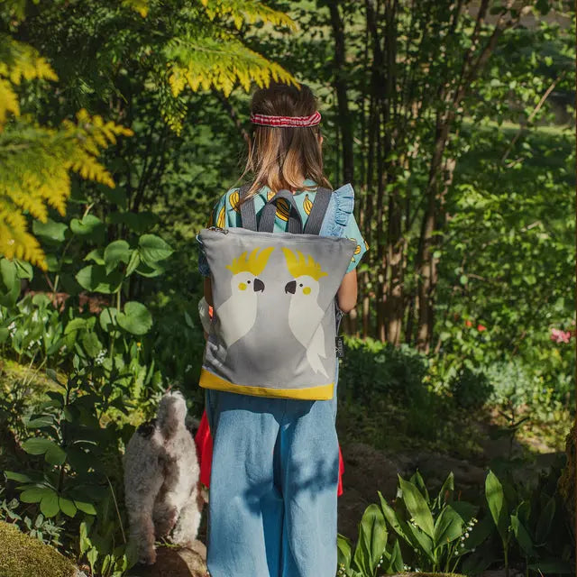 A girl with a backpack and a dog walking in the woods. Kids backpack - Cockatoo: velvet top with Cockatoo design, waterproof polyester bottom, orange lining, beige shoulder straps, zipper closure, 2 slip pockets.