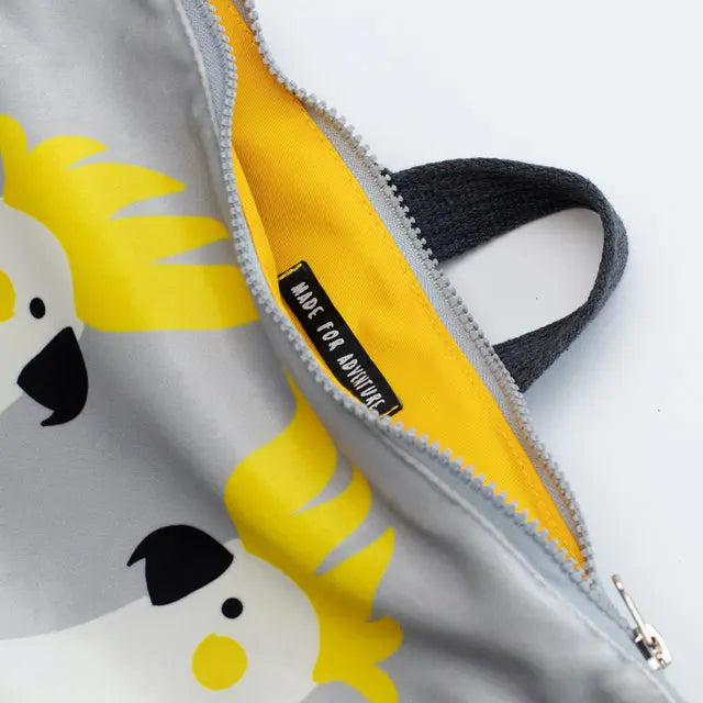 Kids backpack featuring a cartoon dog design, grey with yellow zipper. Lightweight, adjustable straps, velvet top with Cockatoo design, waterproof polyester bottom, orange lining, and two inside slip pockets. Dimensions: 38cm height, 34cm width.