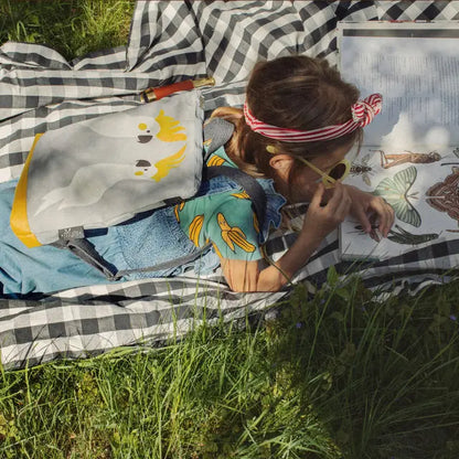 A girl lies on a blanket in the grass, showcasing the Kids backpack - Cockatoo. The backpack features a velvet top with a painted Cockatoo design and sturdy orange polyester bottom. Ideal for 4-10-year-olds.