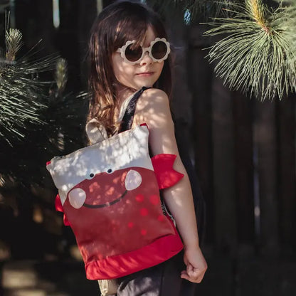 A girl with sunglasses holds a red and white CRAB backpack. Toddler-friendly design for park adventures. Velvet top, waterproof bottom, adjustable straps, and zipper closure. Two sizes available.