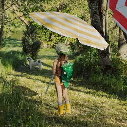 A girl walks with a dog under a yellow and white striped umbrella. A person wears a green backpack. A dog stands in the grass.