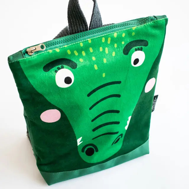 A small kids' backpack featuring a green bag with a crocodile face design. Made of velvet and waterproof polyester, with adjustable straps and two inside pockets. Ideal for ages 2-6.