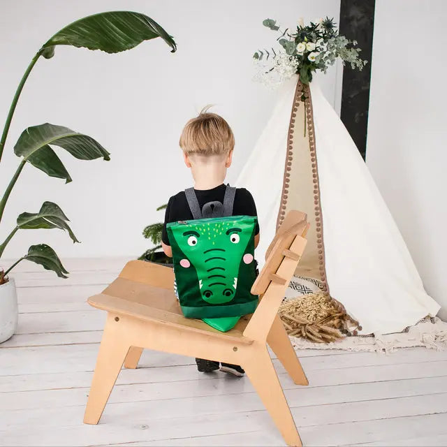 A toddler sits on a chair with a green crocodile backpack. Velvet top with crocodile design, waterproof polyester bottom, adjustable straps, and zipper closure. Ideal for 2-6-year-olds.