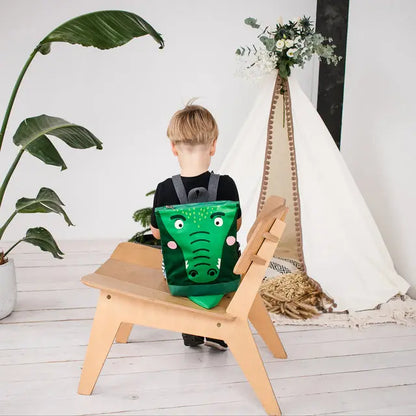 A toddler sits on a chair with a green crocodile backpack. Velvet top with crocodile design, waterproof polyester bottom, adjustable straps, and zipper closure. Ideal for 2-6-year-olds.