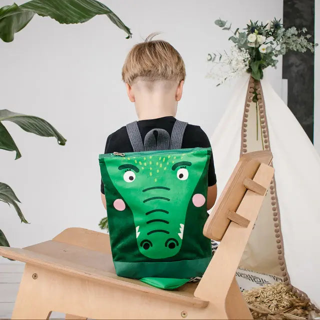 A boy sits on a chair with a green backpack featuring a cute crocodile design. Ideal for ages 2-6, this small backpack is made of velvet and waterproof polyester, with adjustable straps.