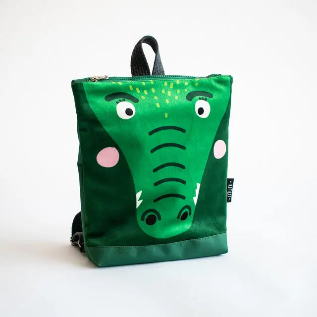 A small green kids backpack featuring a crocodile face design. Made of velvet and waterproof polyester, with adjustable straps and two inside pockets. Ideal for 2-6-year-olds on adventures.