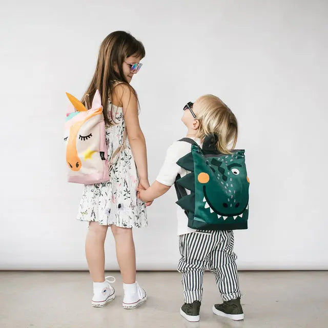 A girl and a boy hold hands, both wearing a Dino-rrific DINOSAUR kids backpack. The backpack features a prehistoric design, velvet top, waterproof polyester bottom, adjustable straps, and zipper closure.