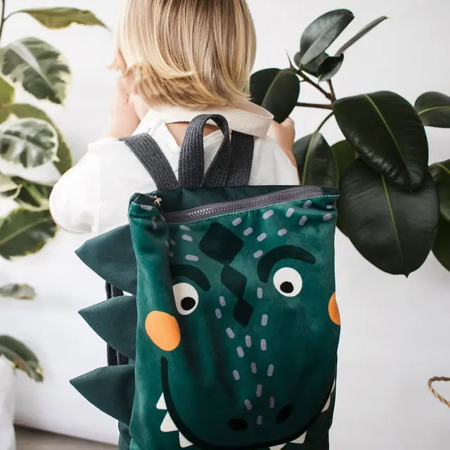 Child holding a green dinosaur kids backpack, featuring prehistoric design, velvet top, waterproof polyester bottom, adjustable straps, and zipper closure. Perfect for young adventurers on outdoor escapades.