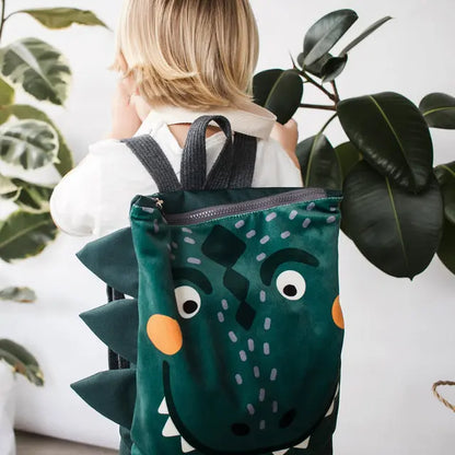 Child holding a green dinosaur kids backpack, featuring prehistoric design, velvet top, waterproof polyester bottom, adjustable straps, and zipper closure. Perfect for young adventurers on outdoor escapades.