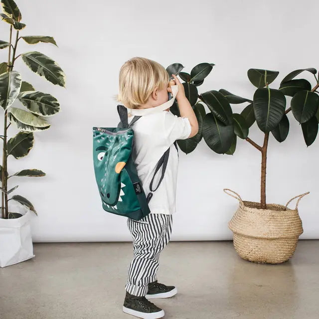 A child with a Dino-rrific DINOSAUR kids backpack, featuring a prehistoric design for outdoor adventures. Made of velvet fabric with a waterproof bottom part in dark green. Two adjustable shoulder straps and zipper closure.