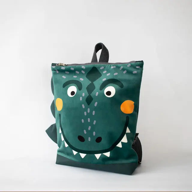 A green kids backpack with a cartoon dinosaur face design, perfect for young adventurers. Features include velvet top with dinosaur print, waterproof polyester bottom, adjustable straps, and zip closure.